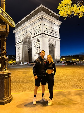 Hombre y mujer de pie frente al Arco del Triunfo iluminado por la noche. Hay un poste con señalización, árboles y vehículos en la calle