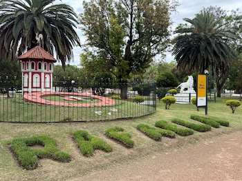 Un reloj en el suelo rodeado por una reja, palmeras, césped con la palabra Rafaela y un cartel amarillo con la leyenda Reloj Floral