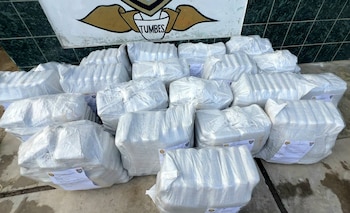 Montones de paquetes rectangulares envueltos en plástico blanco transparente, apilados en el suelo, con el escudo de "Tumbes" al fondo