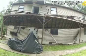 Una casa de dos pisos con terraza de madera parcialmente colapsada en su frente. Una motocicleta con lona negra está en el jardín. Se observa un texto digital con fecha