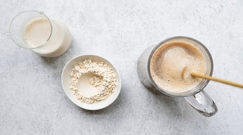 Jarra de vidrio con leche, tazón blanco con avena, vaso de vidrio con bebida espumosa y pajita, sobre superficie de color claro.