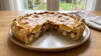 Una tarta dorada sobre un plato de cerámica muestra un corte con relleno de trozos de pollo, cebolla y rodajas de huevo cocido.