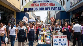 Varias mujeres con delantales ofrecen regalos y herramientas en un mercado callejero. Un arco con el texto 'Mesa Redonda' se ve al fondo.