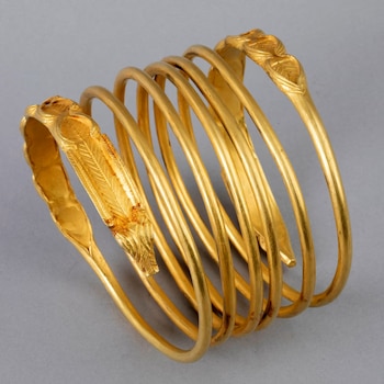 Brazalete de oro antiguo en espiral con siete vueltas y extremos finamente decorados con motivos de hojas y plumas, exhibido sobre un fondo liso