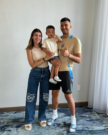 Gerónimo Rulli, vestido con una camiseta dorada de Argentina, posa junto a Rocío Espósito Suárez y un niño. Todos sonríen frente a una pared blanca