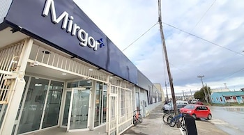 Exterior de un edificio con cartel azul 'Mirgor'. Frente de vidrio, rejas blancas, bicicletas. Calle con autos estacionados, incluyendo uno rojo, bajo cielo nublado