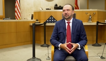 Un hombre con barba, traje azul y corbata roja está sentado en un tribunal. Se ve una bandera de EE.UU. plegada y una estatua de la justicia al fondo