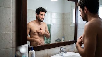 Un hombre sin camiseta se mira en el espejo del baño y coloca los dedos de una mano sobre la zona del pezón, junto al lavabo.