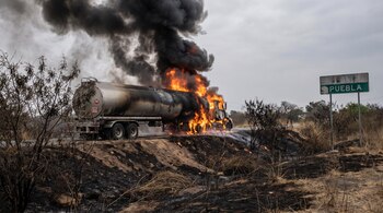 Una pipa de combustible ardiendo en llamas intensas y humo negro denso en una carretera, con vegetación seca y suelo quemado. Se ve un cartel que dice Puebla.