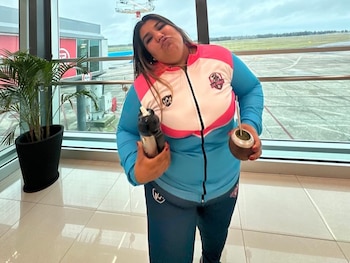 Una mujer joven con un conjunto deportivo azul, rosa y blanco, mate y botella, posa en la terminal de un aeropuerto con la pista de aterrizaje al fondo