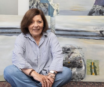 Una mujer de camisa a rayas y pantalones de mezclilla permanece sentada con las piernas cruzadas delante de varias pinturas abstractas