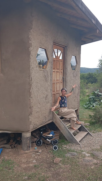 Mujer descalza sonriente sentada en escalones de madera frente a una casa rústica de paredes de tierra. Detrás se ven árboles y colinas verdes