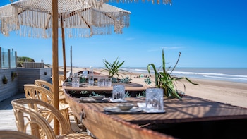 Mesa con vasos y platos bajo una sombrilla de flecos junto a sillas de mimbre, con la arena y el mar de fondo