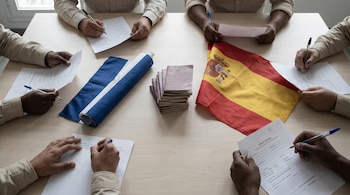 En una mesa de madera, varias manos escriben en formularios. Se ven pasaportes apilados, una bandera de España doblada y una bandera de Nicaragua enrollada.