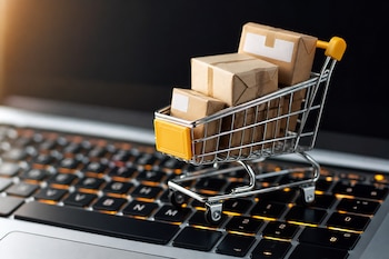 Un carrito de compras metálico en miniatura con cajas envueltas en papel kraft encima de un teclado de computadora retroiluminado.