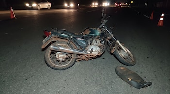 Motocicleta verde oscuro caída en el asfalto por la noche, con asiento separado a un lado. Conos de tráfico naranjas están cerca. Luces de vehículos al fondo.