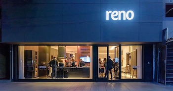 Fachada nocturna de Reno amoblamientos con el cartel iluminado "reno". Grandes ventanales muestran el interior brillante con varias personas