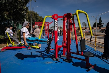 Una mujer de cabello cano realiza ejercicios en una máquina de gimnasio roja y amarilla instalada sobre un suelo azul en un parque