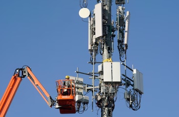 Trabajador en elevador de pluma naranja, montado en una torre de telecomunicaciones con equipos blancos y cables, contra un cielo azul