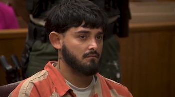 Hombre barbudo con pelo oscuro y uniforme a rayas naranja y blanco, mirando al frente durante una comparecencia judicial, con tatuaje en el cuello