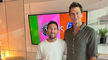 Lionel Messi y Tom Brady posan sonrientes frente a una pantalla que exhibe el logo del Inter Miami, con una planta y libros en el fondo.
