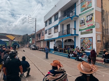 Calle de un pueblo con un grupo de personas reunidas frente a un edificio blanco y azul con el letrero Municipalidad Distrital Huagarancha. Se ven autos y perros