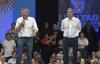 Dos hombres con camisa blanca de pie sobre una tarima, uno habla frente a un micrófono y el público observa al fondo