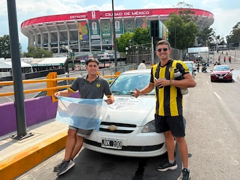 Un auto Chevrolet blanco con calcomanía "América en Corsa" y matrícula argentina está estacionado frente a un estadio con banderas del Mundial 2026