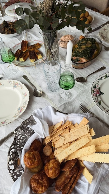 Vista superior de una mesa con mantel blanco, platos con comida, panera con galletas, copas de vidrio verde, florero y cubiertos
