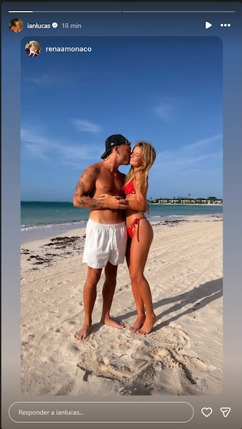 Hombre con tatuajes y bañador blanco besa en la mejilla a mujer con bikini rojo en una playa de arena con el mar y edificios de fondo