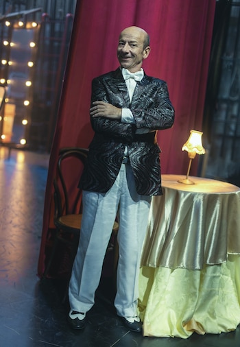 Hombre calvo con chaqueta brillante, pajarita blanca, pantalones blancos y zapatos bicolor. Al fondo, cortina roja, luces borrosas, mesa y lámpara