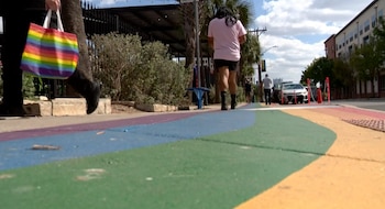 Vista a nivel del suelo de peatones caminando sobre una acera vibrante con franjas de colores azul, verde y amarillo; una persona lleva una bolsa arcoíris