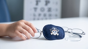 La mano de un niño toca unas gafas con un parche azul marino, junto a una lente suelta y una cartilla optométrica al fondo.