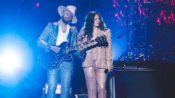 Hombre con sombrero y traje plateado tocando guitarra eléctrica junto a mujer con traje rosa y micrófono, ambos en un escenario con luces azules