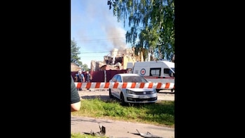Un edificio con daños y humo, una ambulancia blanca, un coche blanco, una barrera de seguridad, personas y árboles