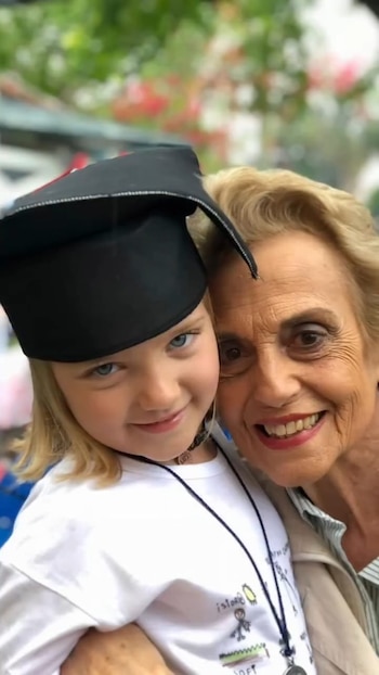 Primer plano de una niña rubia con ojos azules y birrete negro junto a una mujer mayor rubia y sonriente, ambas mirando a cámara