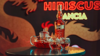 Una botella de aperitivo, una tetera de vidrio con infusión roja y dos vasos pequeños sobre una mesa redonda frente a un letrero luminoso