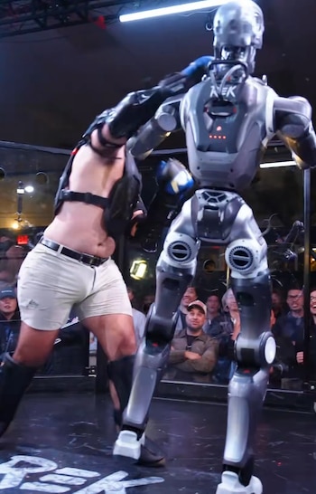 Un robot humanoide de color gris y un hombre con guantes de boxeo y protector pectoral en un cuadrilátero