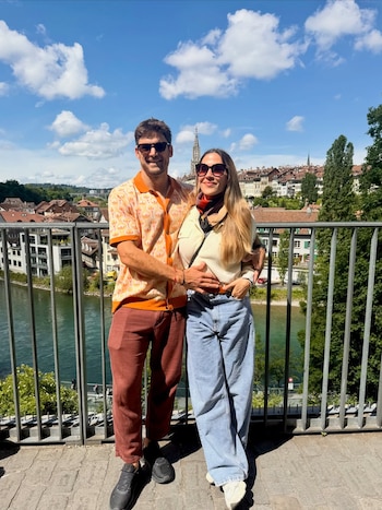 Jimena Barón y Matías Palleiro posan abrazados y sonrientes al aire libre, con gafas de sol, frente a un río, edificios históricos y un cielo con nubes