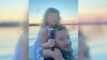 Un hombre lleva a una niña sobre sus hombros. La niña sostiene una linterna encendida frente a un cuerpo de agua al atardecer