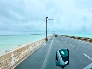 Carretera gris mojada con líneas blancas y un muro de hormigón. A la izquierda, mar turquesa y cielo nublado. Un coche negro se aleja. Espejo retrovisor a la derecha