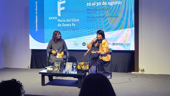 Dos mujeres sentadas en un escenario frente a una pantalla azul sobre la Feria del Libro de Santa Fe, con libros expuestos en una mesa