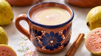 Taza de barro con flores azules llena de atole humeante, junto a guayabas enteras, mitades de fruta y una vara de canela.