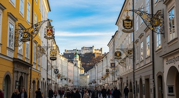 Una calle estrecha con edificios de fachadas claras y letreros ornamentados muestra personas caminando; al fondo, una fortaleza en una colina.
