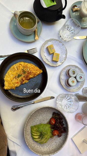 Vista cenital de una mesa con desayuno que incluye un omelet, aguacate rebanado, lechuga, tomates cherry, té, agua, mantequilla y mermeladas