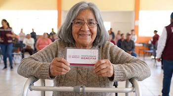 Mujer de cabello gris y gafas sonríe a cámara, sentada detrás de un andador, y muestra una tarjeta blanca del Gobierno de México 'Tarjeta para el Bienestar'.