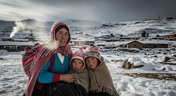 Una mujer andina y dos niños, abrigados con gorros y mantas, sentados en un paisaje nevado con casas rurales y montañas cubiertas de nieve al fondo.