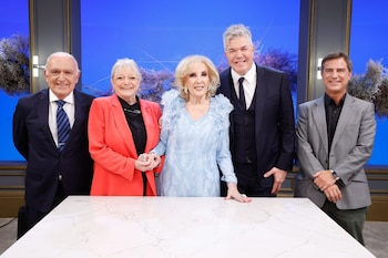 Cinco personas sonrientes, tres hombres y dos mujeres, posan de pie detrás de una mesa blanca frente a un fondo azul con arreglos florales
