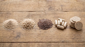 Cinco montones de psyllium en diferentes formatos: polvo, cáscara, semillas, cápsulas y obleas, dispuestos en línea sobre una mesa de madera.