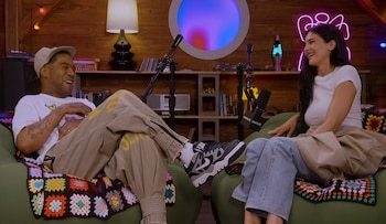 Dos personas sentadas en sillones con mantas de crochet. Un hombre y una mujer, con micrófonos y un fondo de madera con estantes y luces